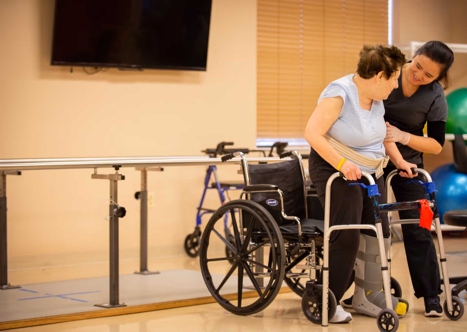 An elderly woman using a walker with a support belt around her waist is assisted by a caregiver in a rehabilitation room. There is a wheelchair nearby, parallel bars for walking exercises, and exercise balls in the background. The room has beige walls and a window with closed blinds.
