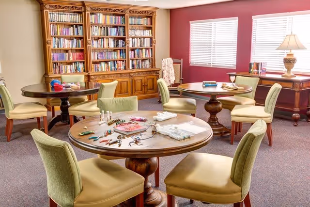 A cozy activity room with round wooden tables and green cushioned chairs. One table has various craft supplies and tools spread out. There is a large wooden bookshelf filled with books against the wall, and a window with white blinds letting in natural light. A wooden side table with a lamp and some books is also visible.