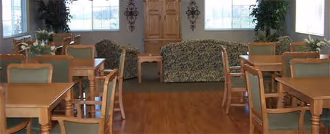 A spacious room with wooden floors featuring multiple wooden tables and chairs arranged for dining. At the far end, there are two patterned sofas and a wooden cabinet against the wall, with large windows letting in natural light and green plants placed near the windows.