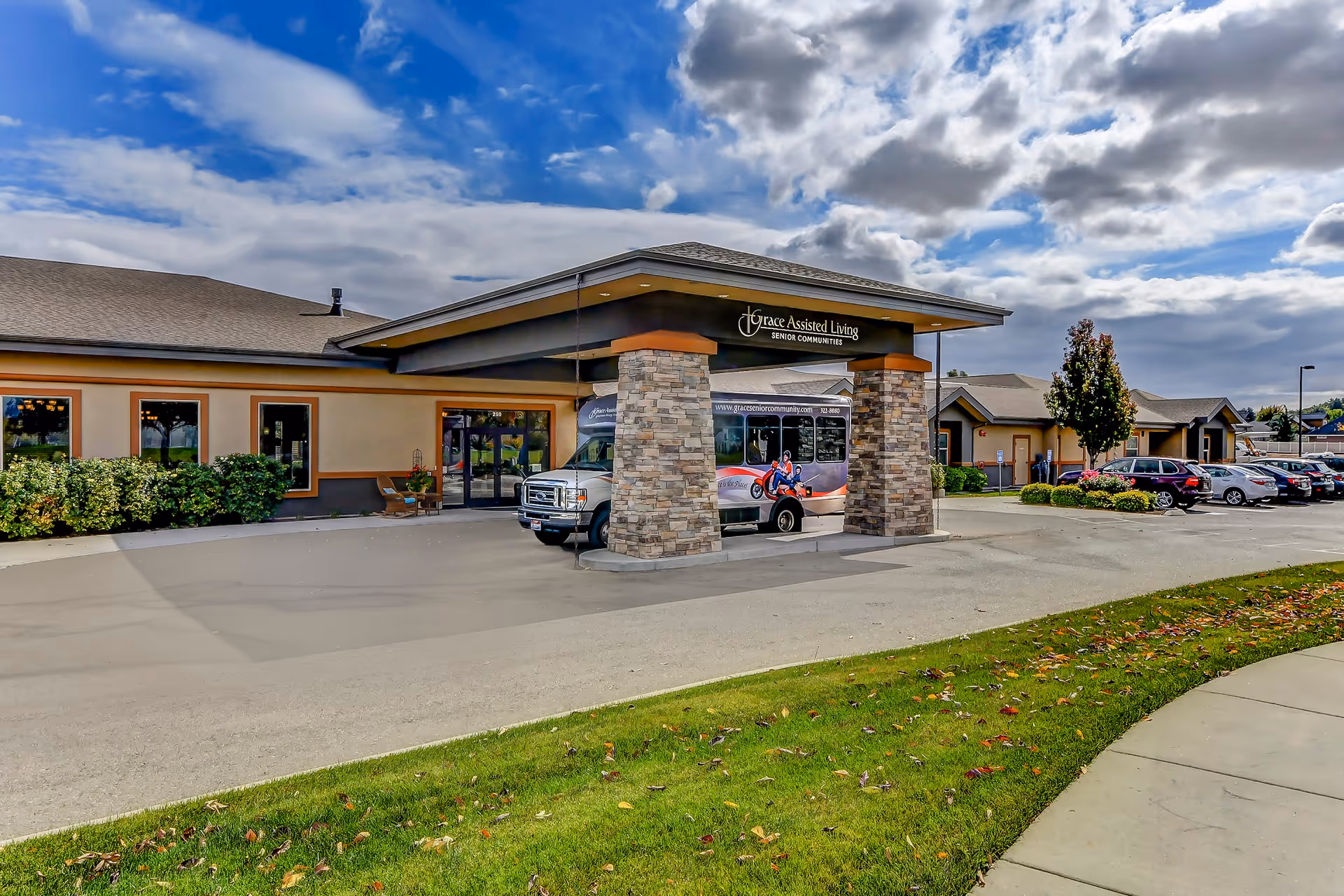 Exterior view of Grace Assisted Living facility showing the main entrance with a covered drop-off area supported by stone pillars. A shuttle bus is parked under the entrance canopy. The building is surrounded by a parking lot with several cars and landscaped greenery. The sky is partly cloudy.