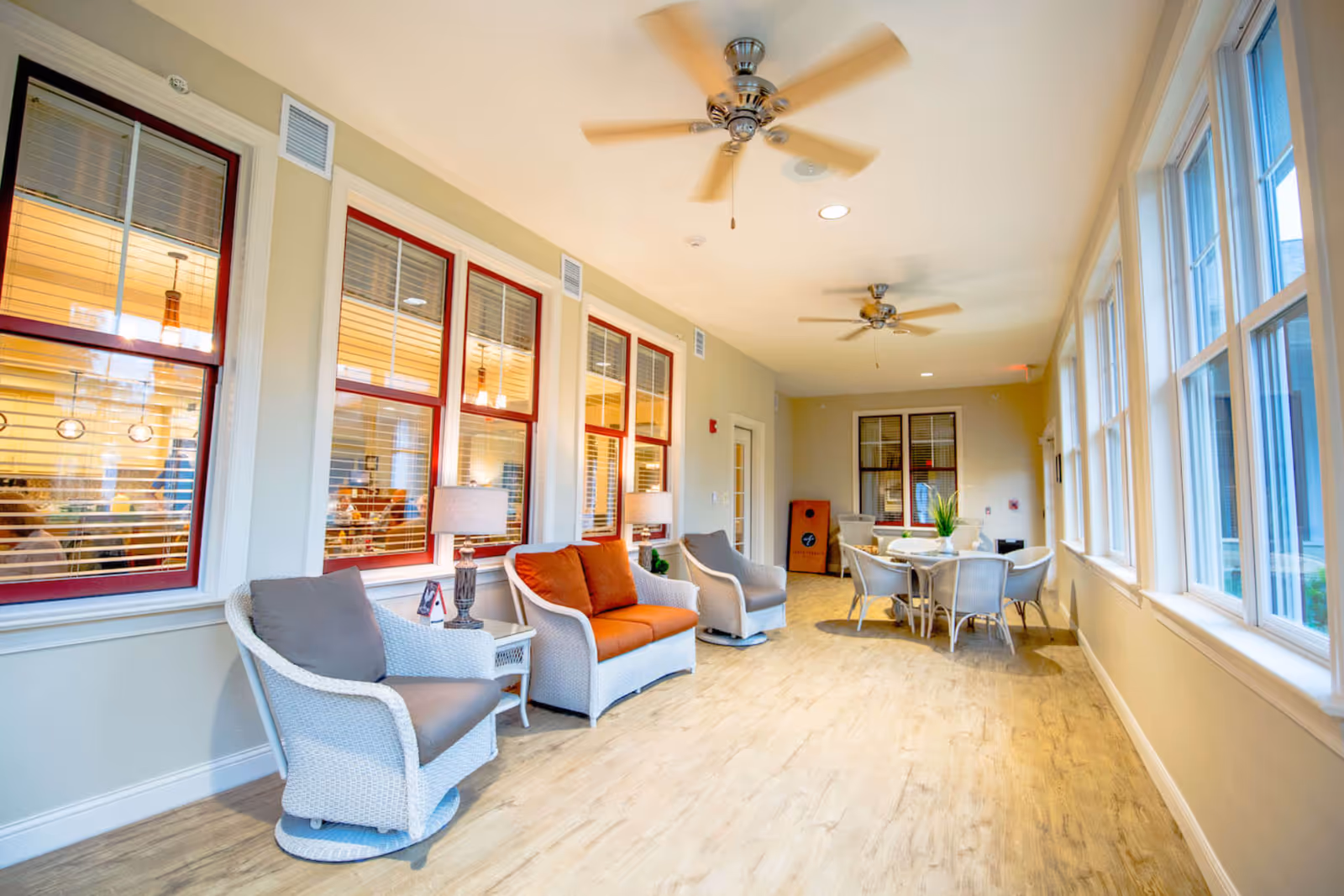 A bright and spacious indoor seating area with wicker chairs and a loveseat with orange cushions, two table lamps, ceiling fans, and a round table with chairs near large windows letting in natural light.