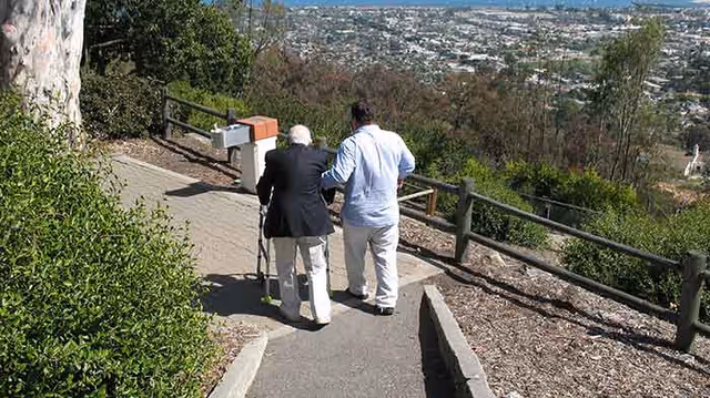 An elderly person using a walker is being assisted by a caregiver as they walk down a paved outdoor path with railings and greenery on the sides, overlooking a cityscape in the background.