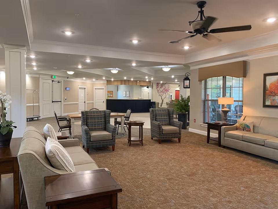 A spacious and well-lit senior living facility common area with beige walls and carpeted floor. The room features two beige sofas, two plaid armchairs, wooden side tables, a ceiling fan, and a large window with blinds. In the background, there is a reception desk and a hallway with doors. The area is decorated with plants, a painting, and a wall decal of a tree.