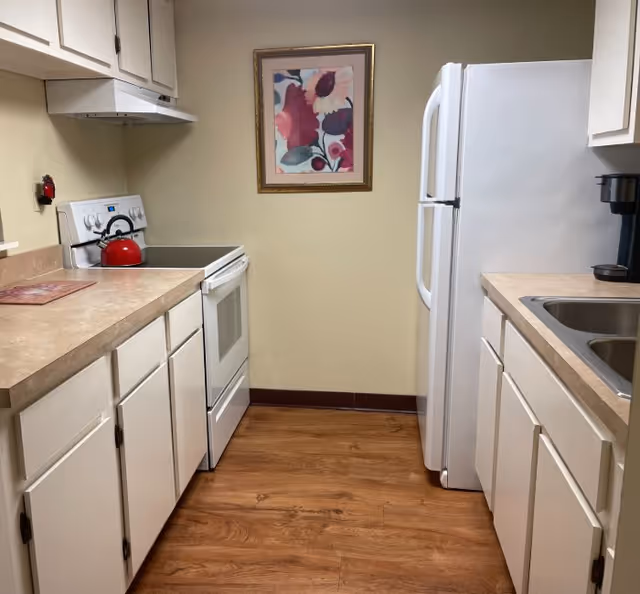 A small kitchen with white cabinets and beige countertops on both sides. On the left side, there is a white electric stove with a red kettle on top and a range hood above it. On the right side, there is a white refrigerator and a sink with a black coffee maker next to it. A framed floral painting hangs on the beige wall at the end of the kitchen. The floor is wood-patterned.