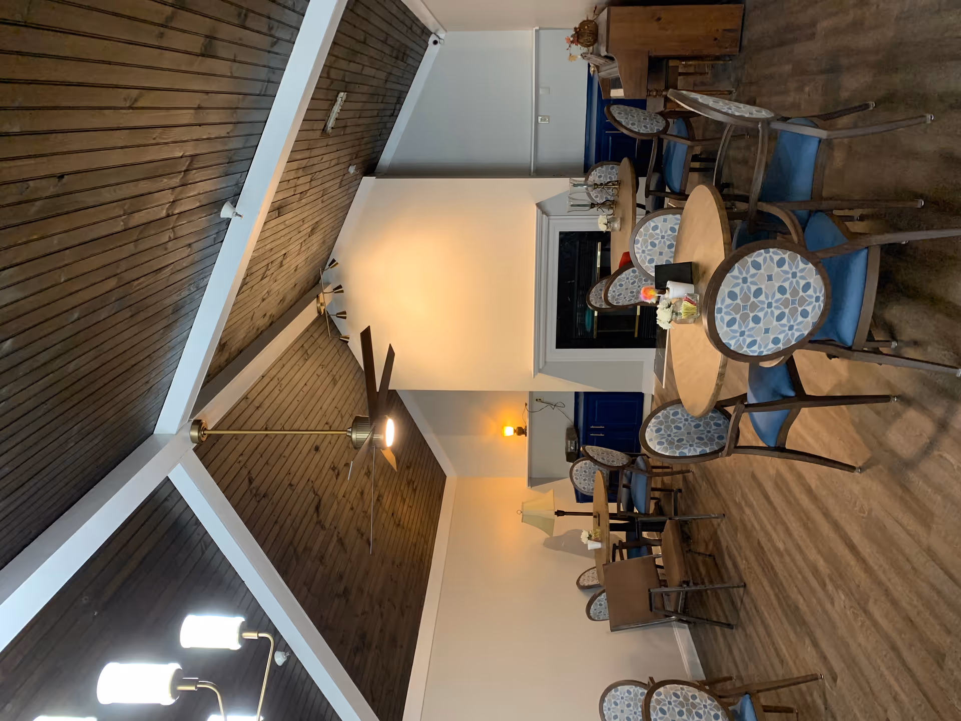 Interior of a senior living facility common area with round wooden tables and blue cushioned chairs arranged around them. The room features a high wooden ceiling with white beams, a ceiling fan, a chandelier, a fireplace, and a piano in the corner. There are small decorative items on the tables and shelves.