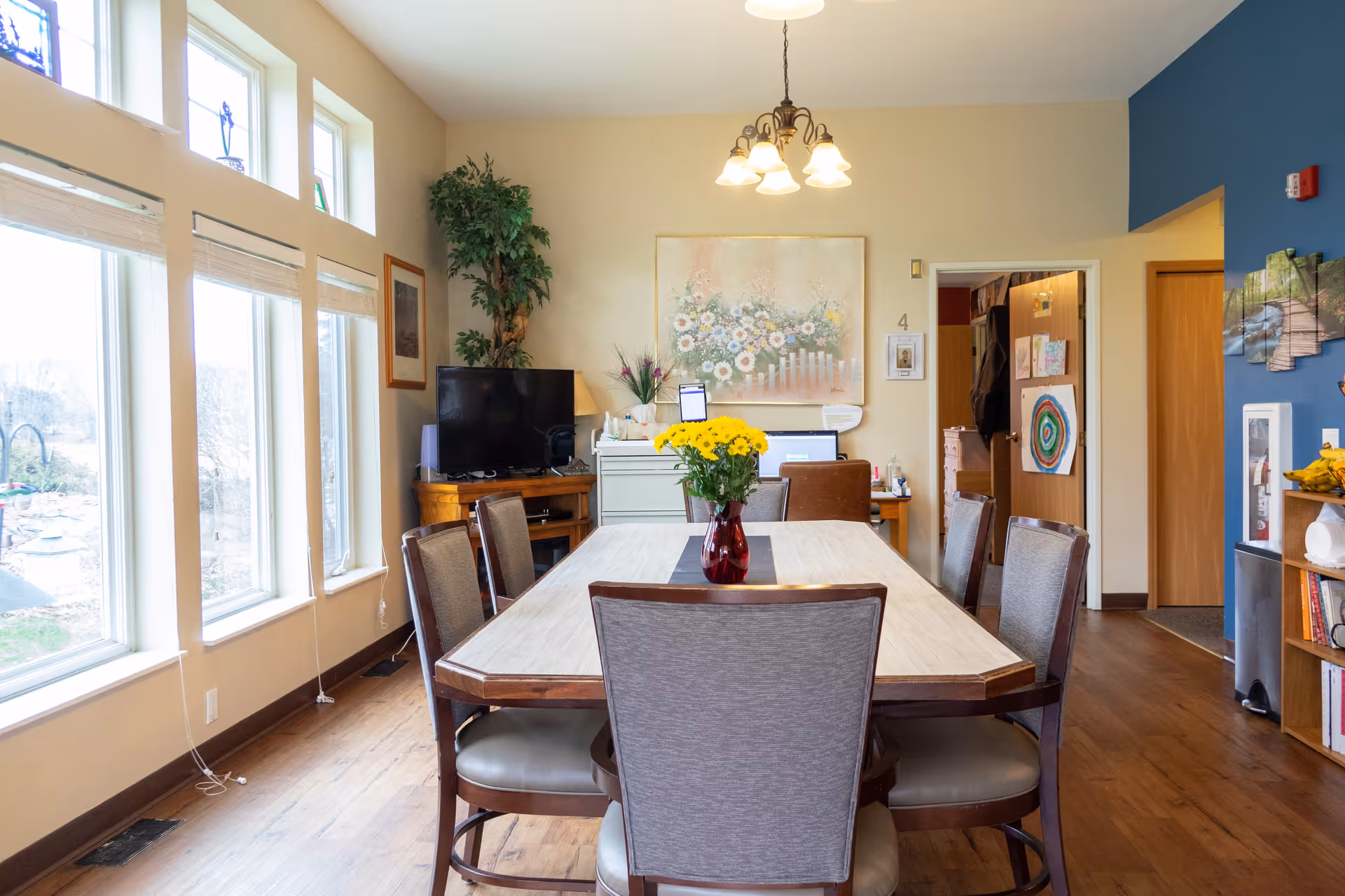 A bright dining room with a rectangular wooden table surrounded by six upholstered chairs. A vase with yellow flowers is centered on the table. Large windows on the left side let in natural light. The room has wooden flooring, a TV on a wooden stand, a potted plant, and various wall decorations including a large floral painting and children's artwork. A chandelier with five lights hangs above the table.