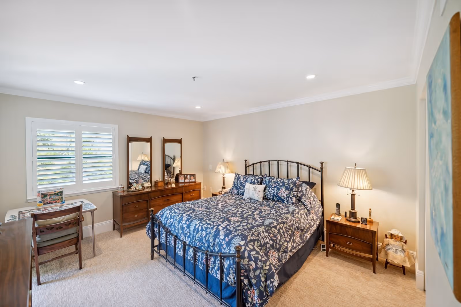A cozy bedroom with a large bed featuring a dark metal frame and blue floral bedding. There are two wooden nightstands with lamps on either side of the bed. A wooden dresser with two mirrors and various decorative items is against the wall. A small table with a chair is positioned near a window with white shutters. The room has beige walls and carpeted flooring.