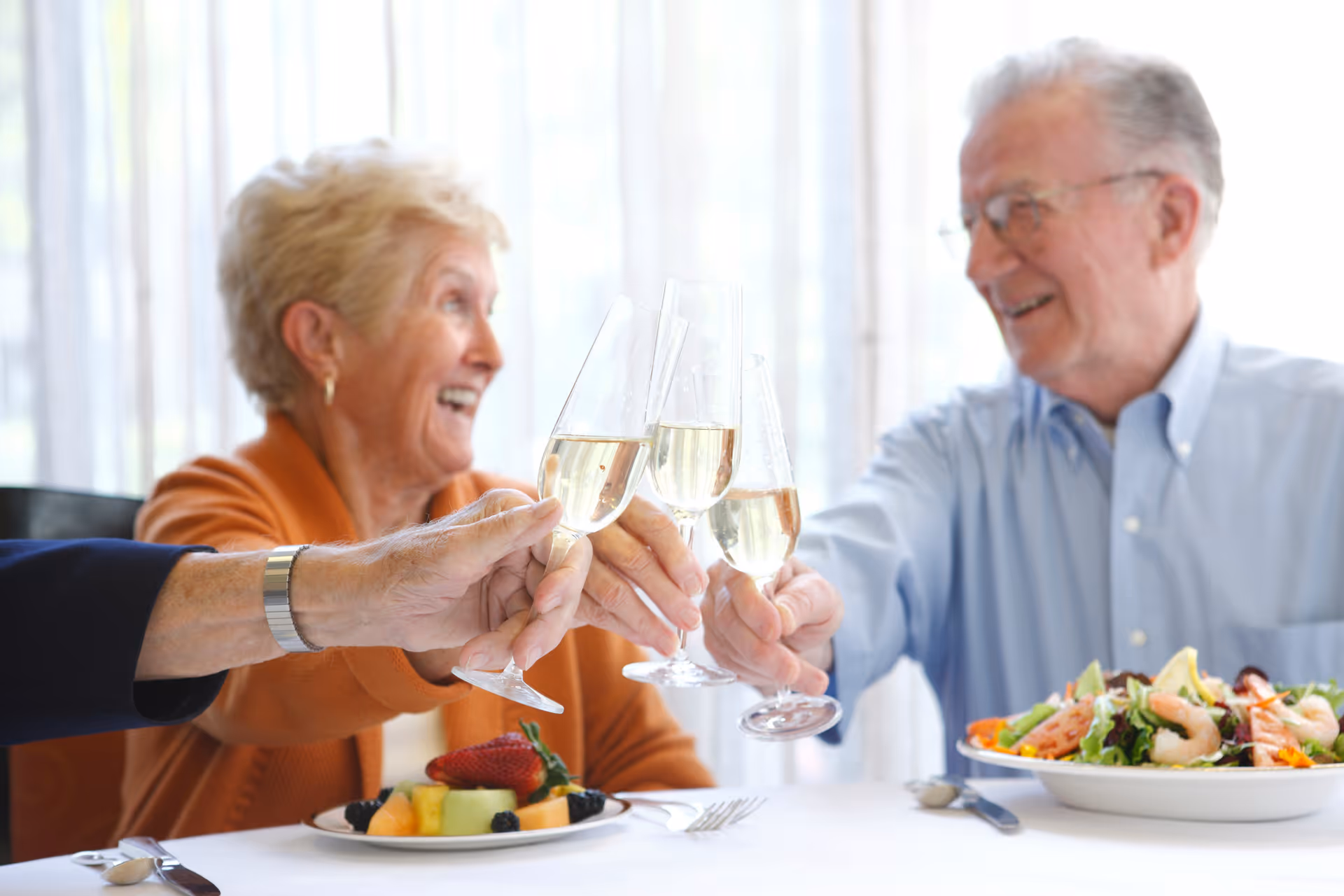 Three older adults toast with champagne flutes over a table set with fruit and a salad.