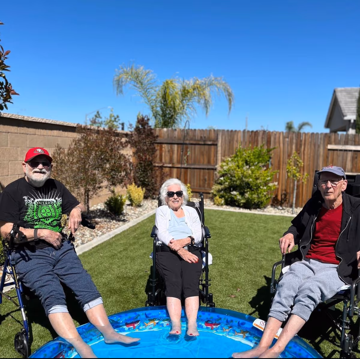 Three elderly individuals sitting in wheelchairs with their feet in a small inflatable pool in a sunny backyard with green grass, plants, and a wooden fence.