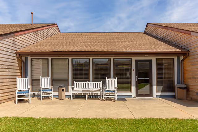 Outdoor patio area of a senior living facility with a concrete walkway, green grass in the foreground, and a building with wooden siding and a shingled roof. There are two white rocking chairs and a white bench with blue cushions arranged along the wall, with a trash can placed between the chairs and bench. The building has large windows with blinds and a glass door.