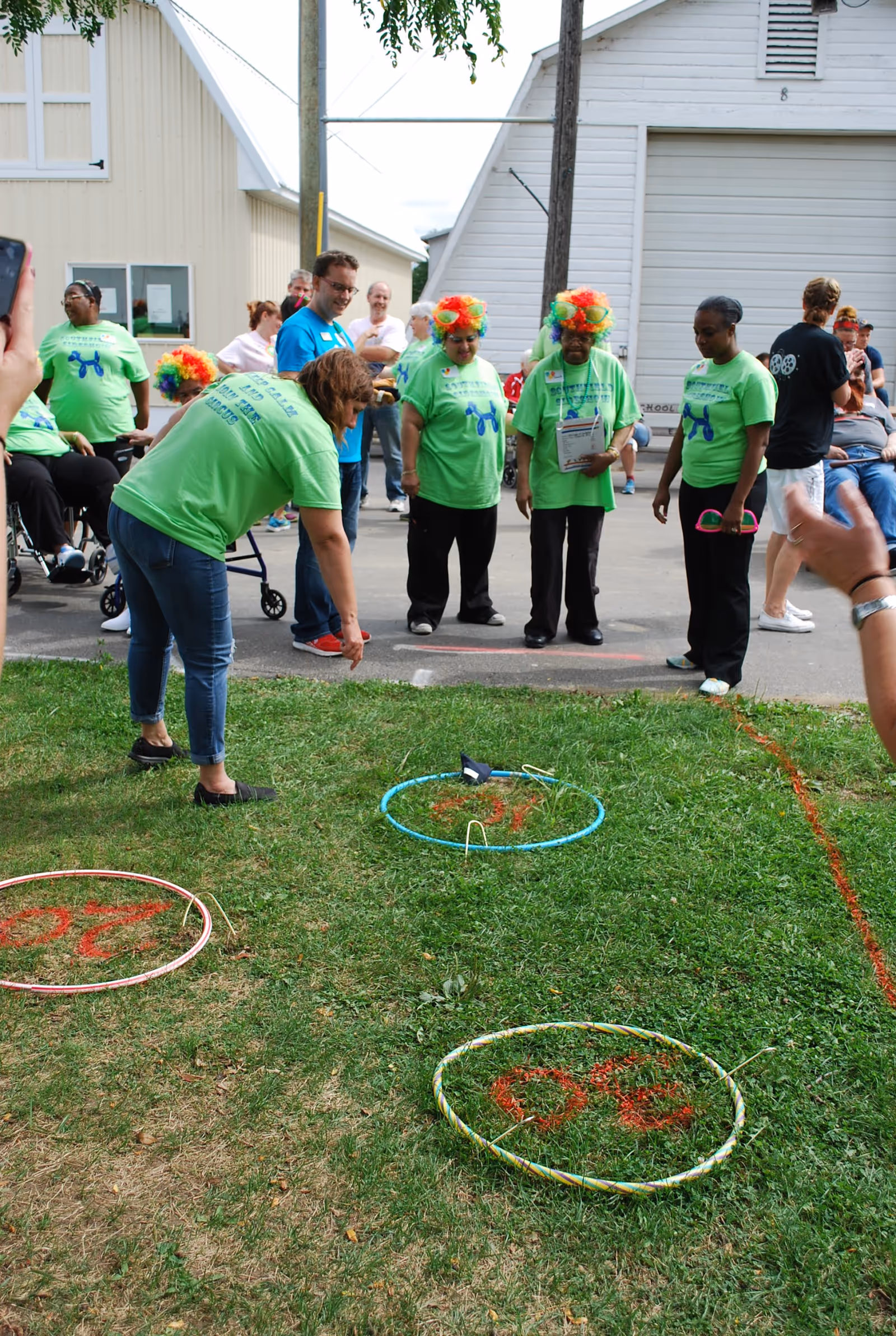 A group of people, some wearing bright green shirts and colorful clown wigs, are gathered outdoors on a grassy area near white buildings. One person is bending down, appearing to participate in a ring toss game with hula hoops placed on the grass marked with numbers. Other people are standing or sitting nearby, watching the activity.