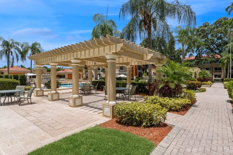 Outdoor courtyard with a shaded pergola, tables and chairs, palm trees and a pool in the background.