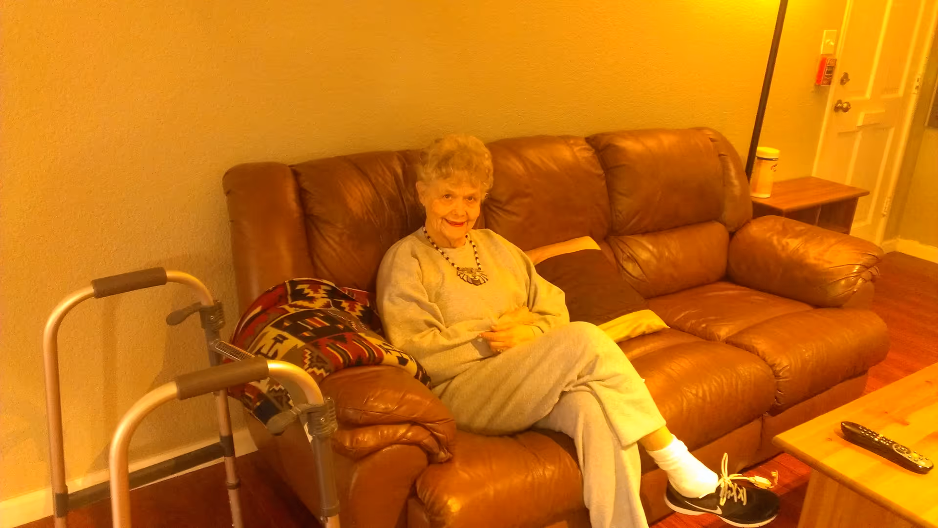 An elderly woman sits on a brown leather sofa in a living room with a walker beside her and a coffee table with a remote in front.