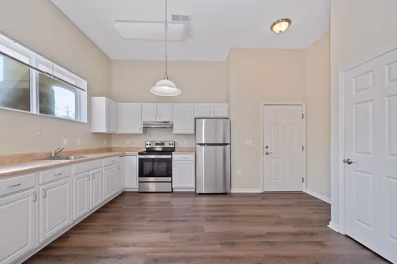 A modern kitchen with white cabinets, a stainless steel refrigerator and oven, a beige countertop, a window above the sink, and a hanging light fixture. The floor is wood with a medium brown finish, and there are two white doors visible on the right side of the image.