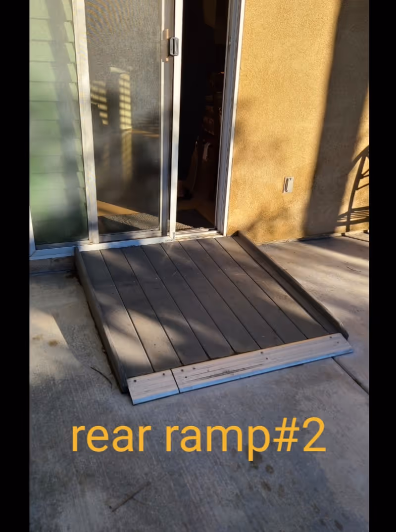A small wooden ramp leading up to a sliding glass door entrance with a screen door, attached to a building with a yellowish exterior wall. The ramp is placed on a concrete surface and there is text at the bottom of the image that reads 'rear ramp#2'.