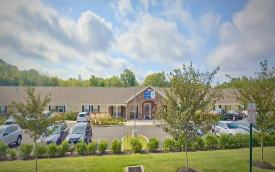 Exterior view of a single-story senior living facility building with a parking lot in front. The building has a gable roof and a central entrance with a small covered porch. Several cars are parked in the lot, and there are small trees and shrubs in landscaped areas near the parking spaces. The sky is partly cloudy.