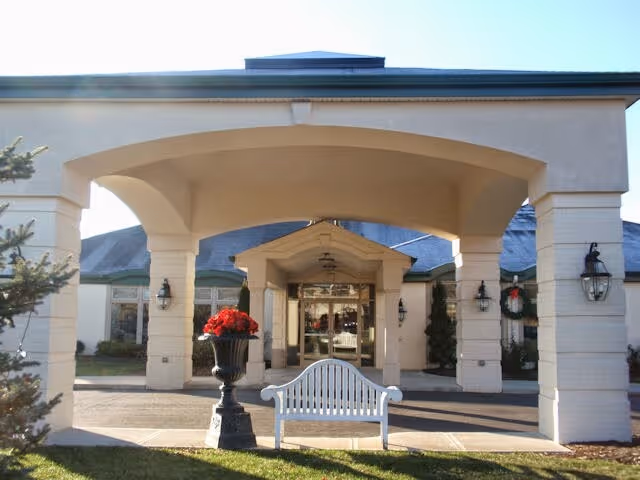 Entrance of a building with a covered driveway supported by large columns. There is a white bench and a large planter with red flowers in front of the entrance. The building has a light-colored exterior with several windows and outdoor wall lanterns.