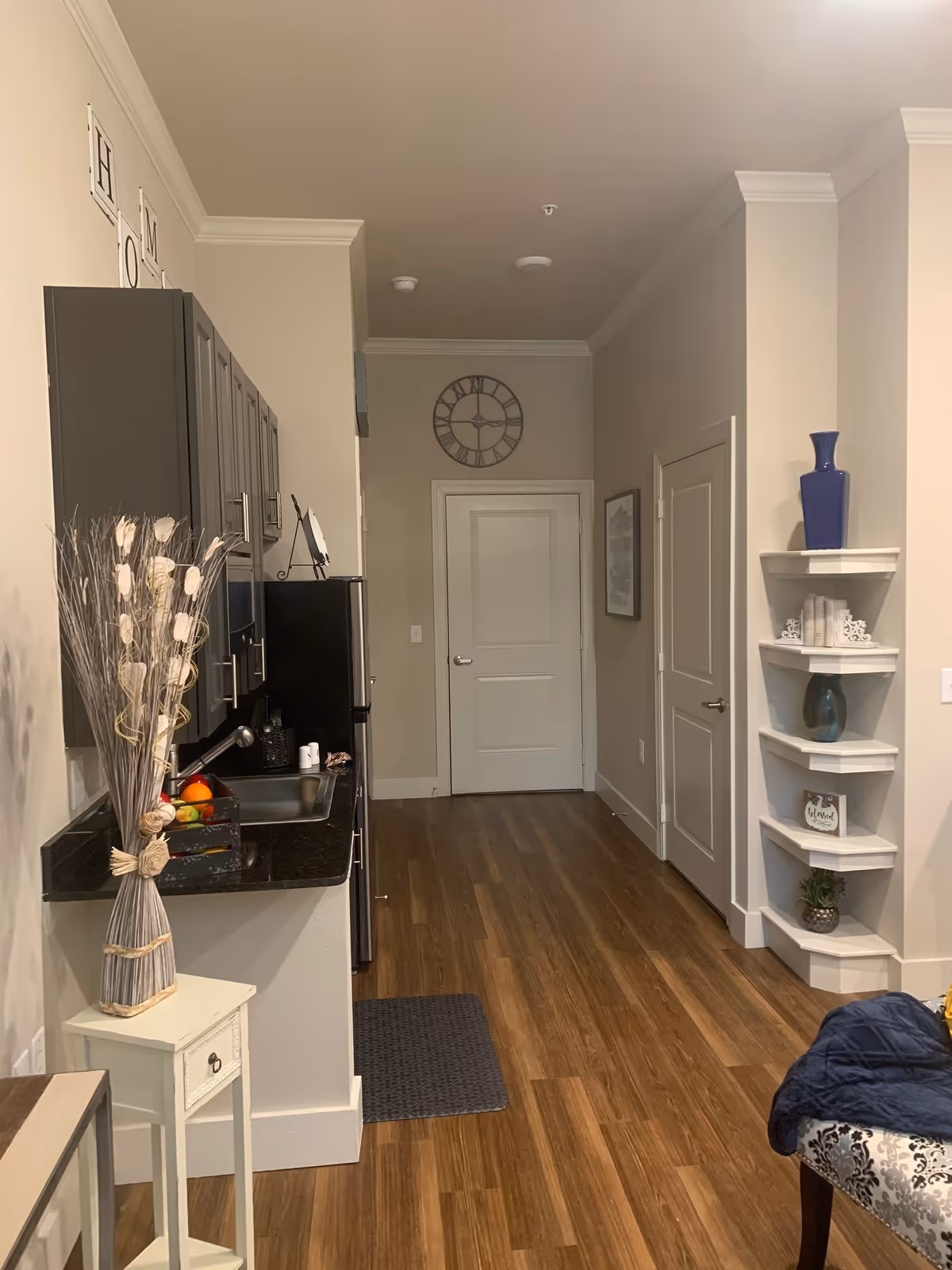 Interior view of a hallway in an assisted living facility with wooden flooring, a small kitchen area on the left with dark cabinets and a black countertop, a stainless steel refrigerator, a wall clock above a white door at the end of the hallway, and corner shelves on the right holding decorative items including a blue vase and small plants.