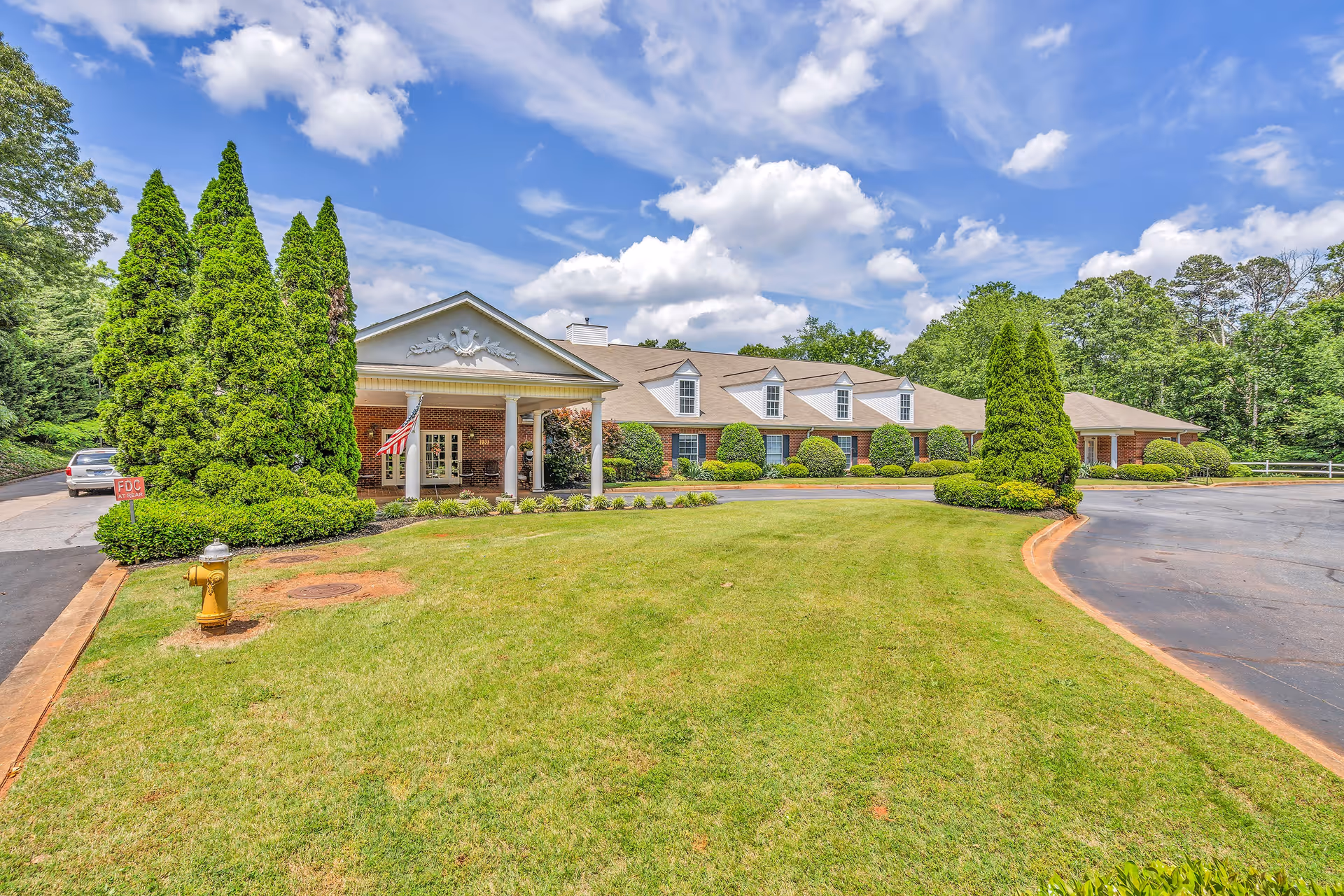 Front exterior view of Windsor House Greenville, a single-story brick building with a covered entrance, surrounded by neatly trimmed bushes, tall evergreen trees, and a well-maintained lawn under a partly cloudy blue sky.