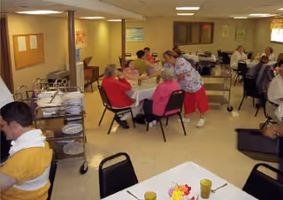 A communal dining area in an assisted living facility with several elderly residents seated at tables. A caregiver is interacting with one of the residents. The room has simple decor with bulletin boards on the walls and a serving cart with plates and utensils.