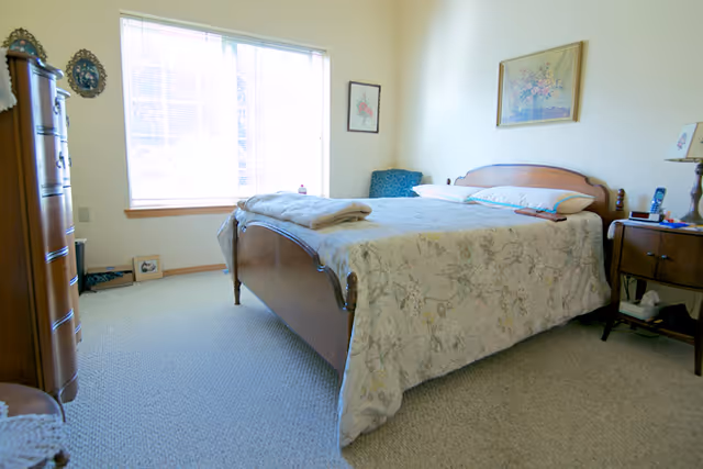 Sunlit bedroom with a wooden sleigh bed covered in a floral bedspread, a large window to the left, and wooden nightstands and dresser.
