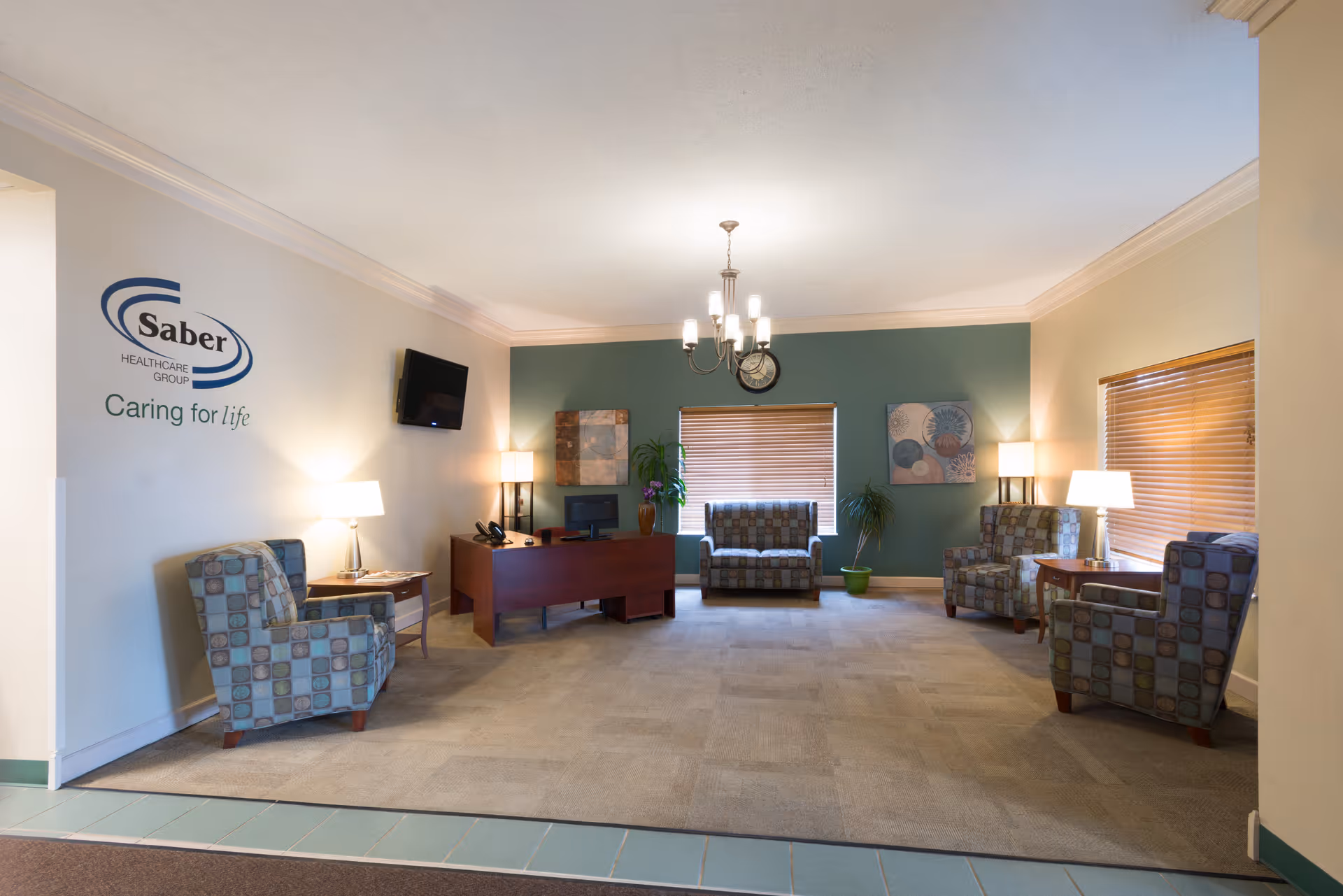 A well-lit living room area with patterned armchairs arranged around the room, a wooden desk with a computer and phone, two windows with wooden blinds, two table lamps, two floor lamps, a wall-mounted TV, and decorative wall art. The wall on the left features the Saber Healthcare Group logo and the phrase 'Caring for life.'