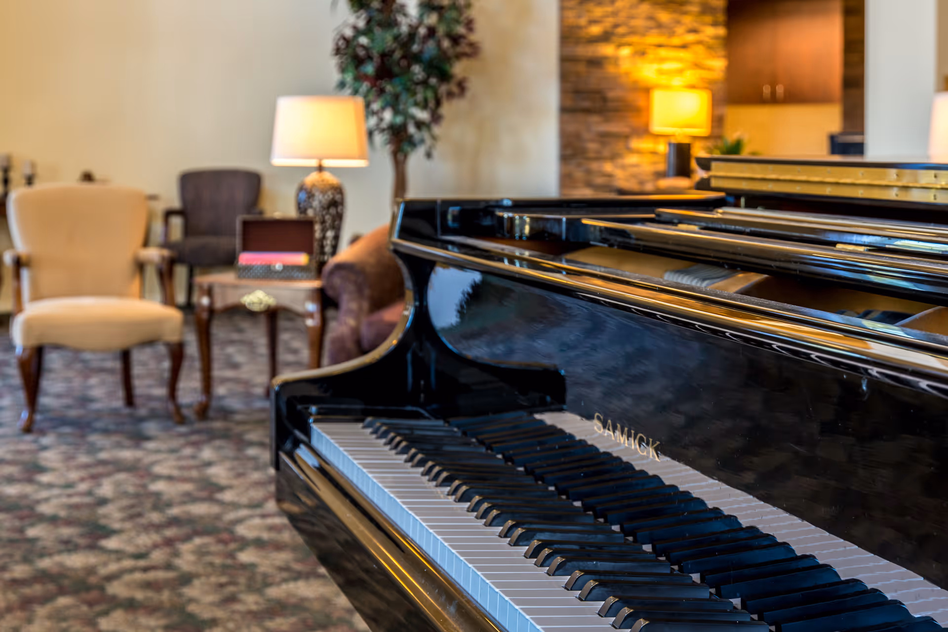 A black grand piano in a warm communal lounge with upholstered chairs, side tables, and lamps.