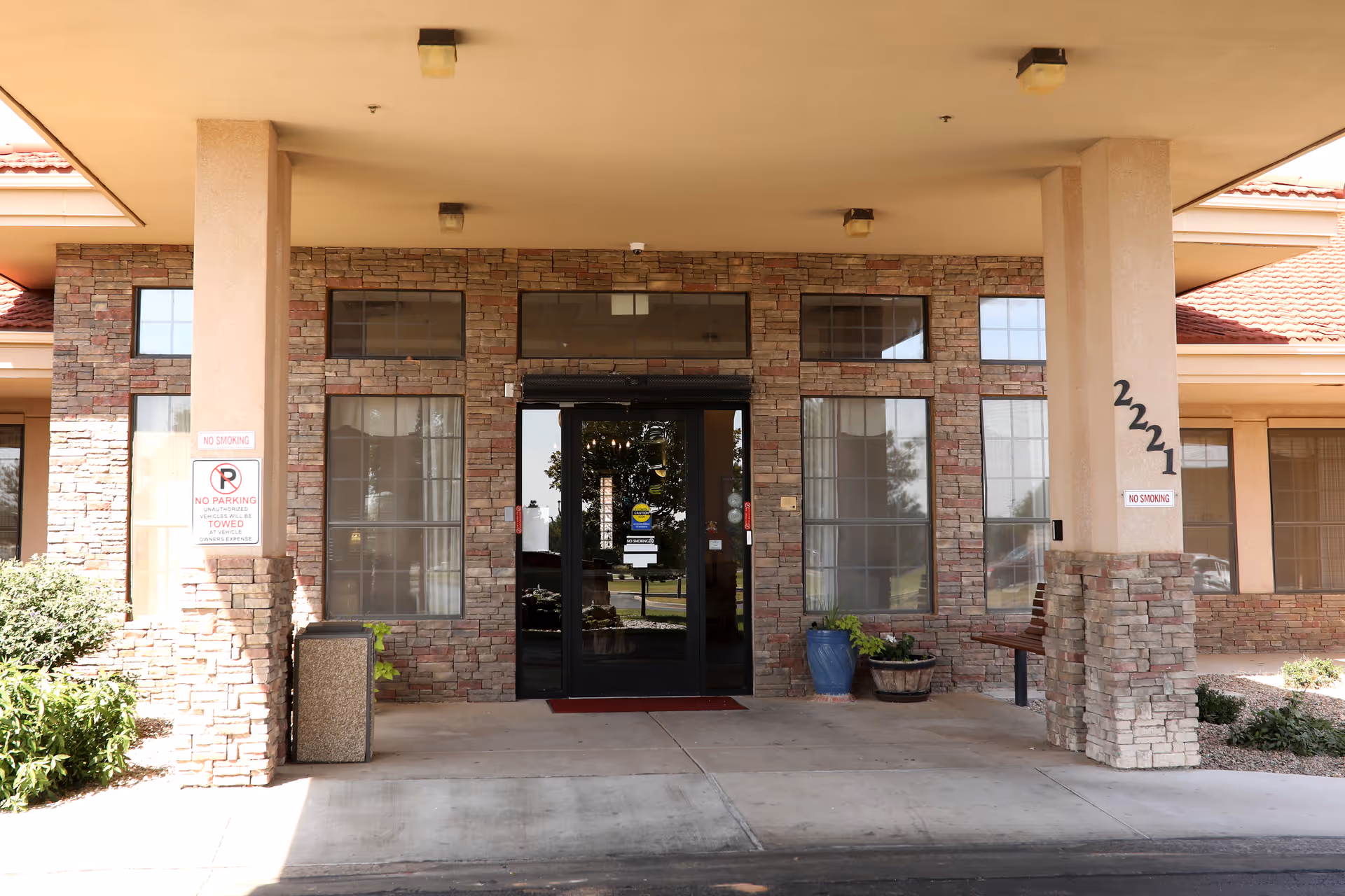 Entrance of a building with stone and brick facade, glass double doors, two large windows on each side, and two columns supporting a roof overhang. There are signs indicating no smoking and no parking, a trash bin, a bench, and potted plants near the entrance.