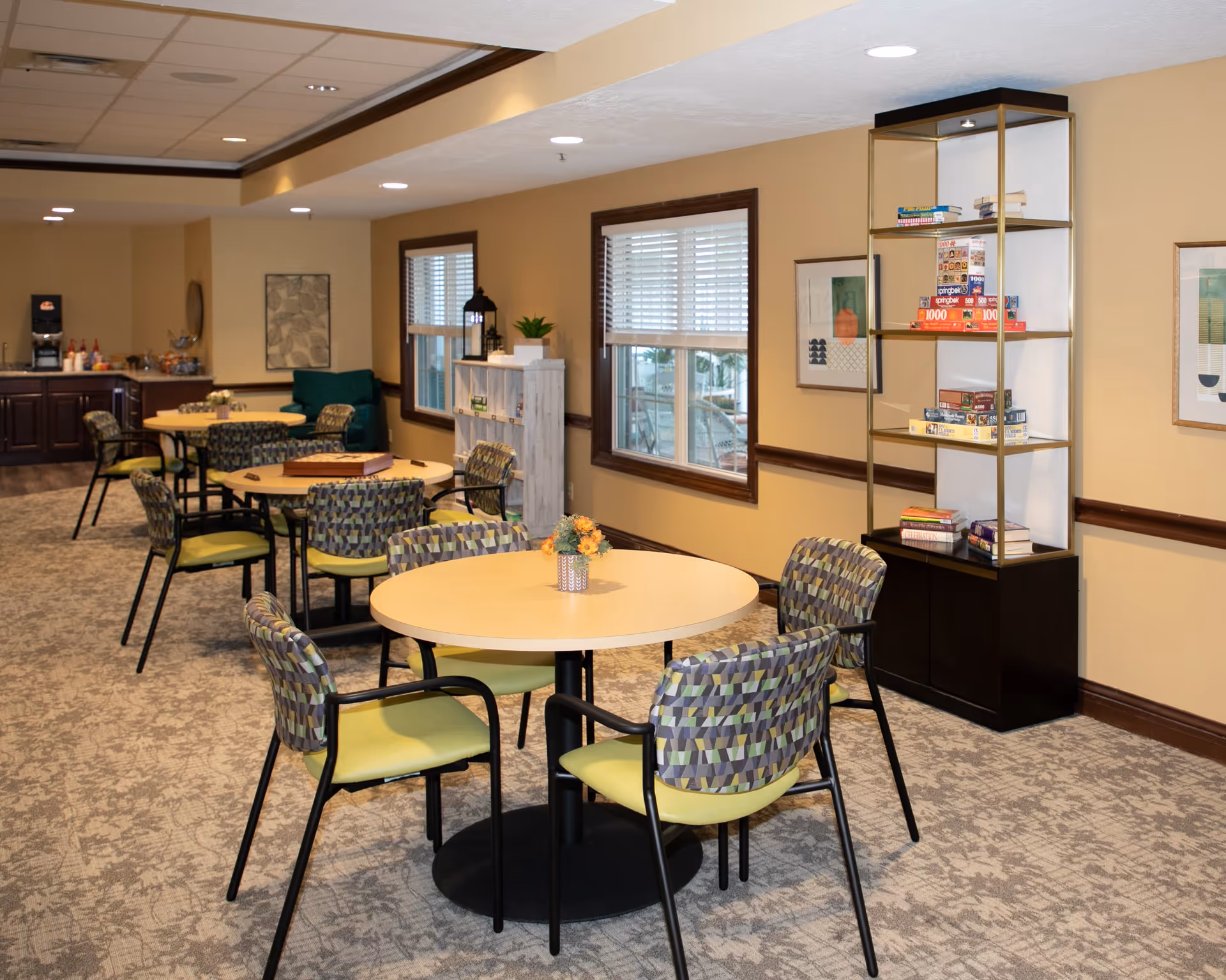 A well-lit common area with round tables and patterned chairs arranged for seating. The room has beige walls with two windows featuring white blinds. There is a tall glass display cabinet filled with board games and books, and a small white shelf with decorative items. In the background, there is a counter with a coffee machine and condiments.