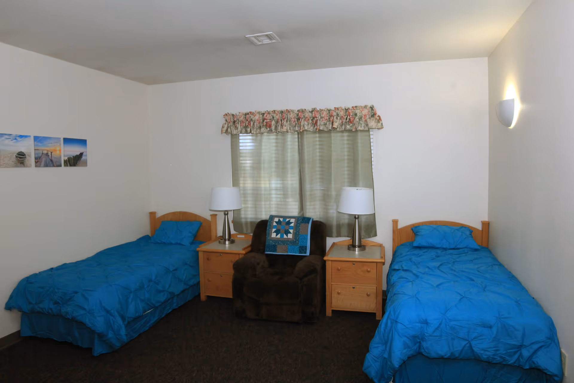A bedroom with two single beds covered in blue bedding, each with a pillow. Between the beds are two wooden nightstands, each with a lamp. In the center, there is a dark brown armchair with a blue and white quilt draped over the back. A window with green curtains and a floral valance is behind the chair. Three small landscape pictures are hung on the left wall.