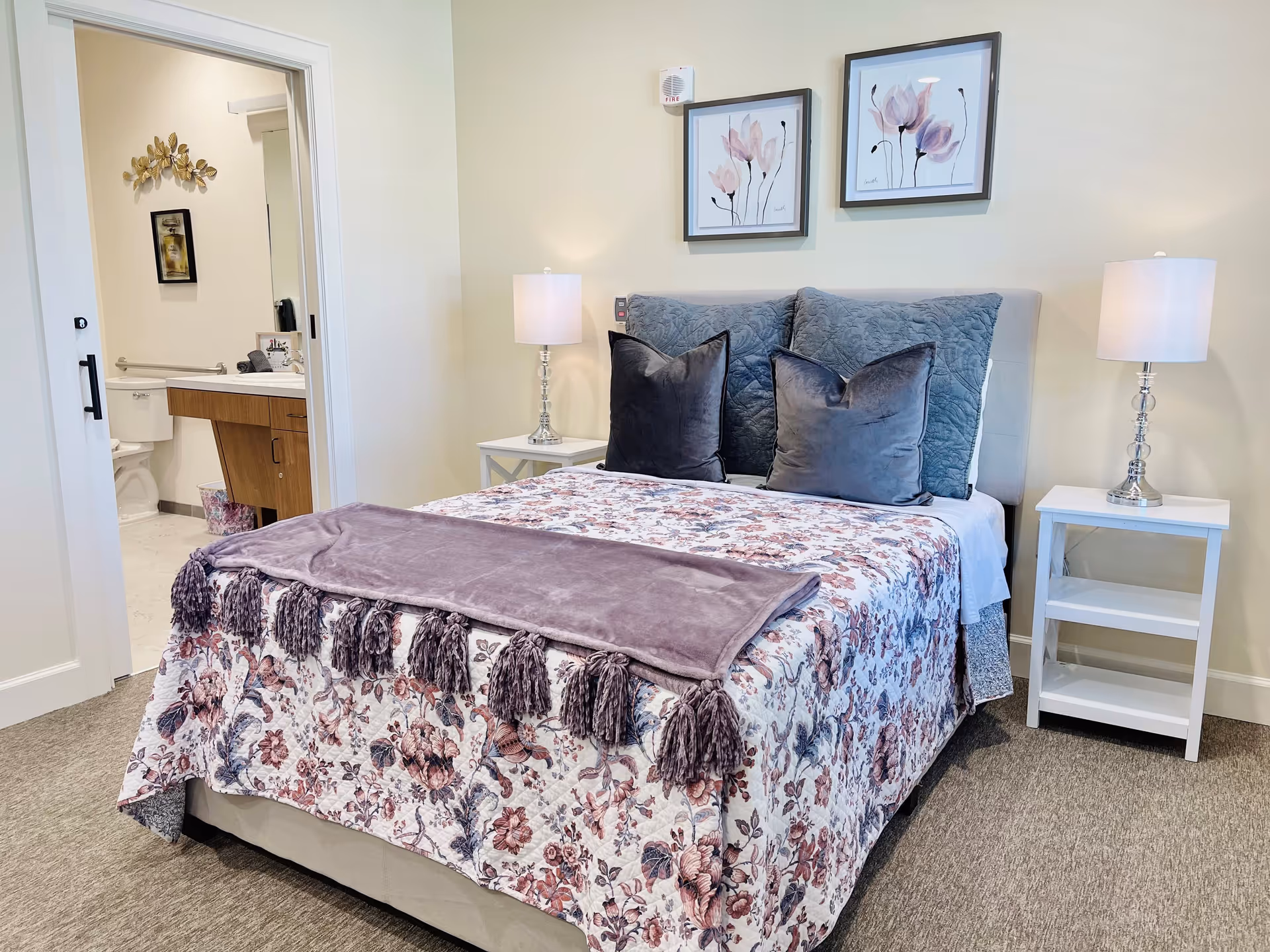A cozy bedroom with a bed covered in a floral quilt and a purple throw blanket. The bed has two large gray pillows and two smaller dark gray pillows. On each side of the bed are white nightstands with silver lamps. Two framed floral artworks hang on the wall above the bed. An open door reveals a bathroom with a toilet, sink, and wall decorations.