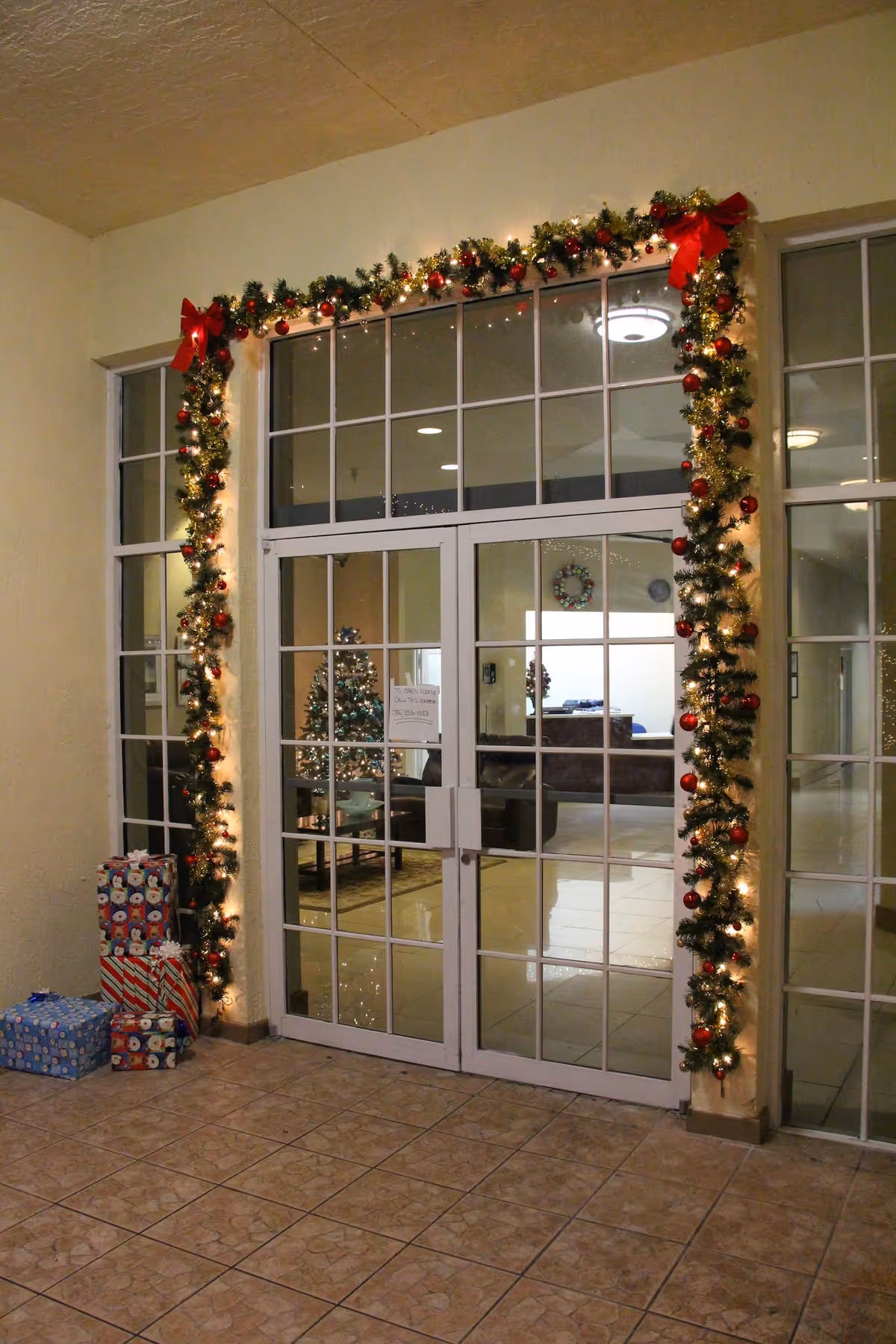 Glass double doors framed by lit Christmas garlands and bows, wrapped presents on the floor, and a decorated tree visible through the doors.
