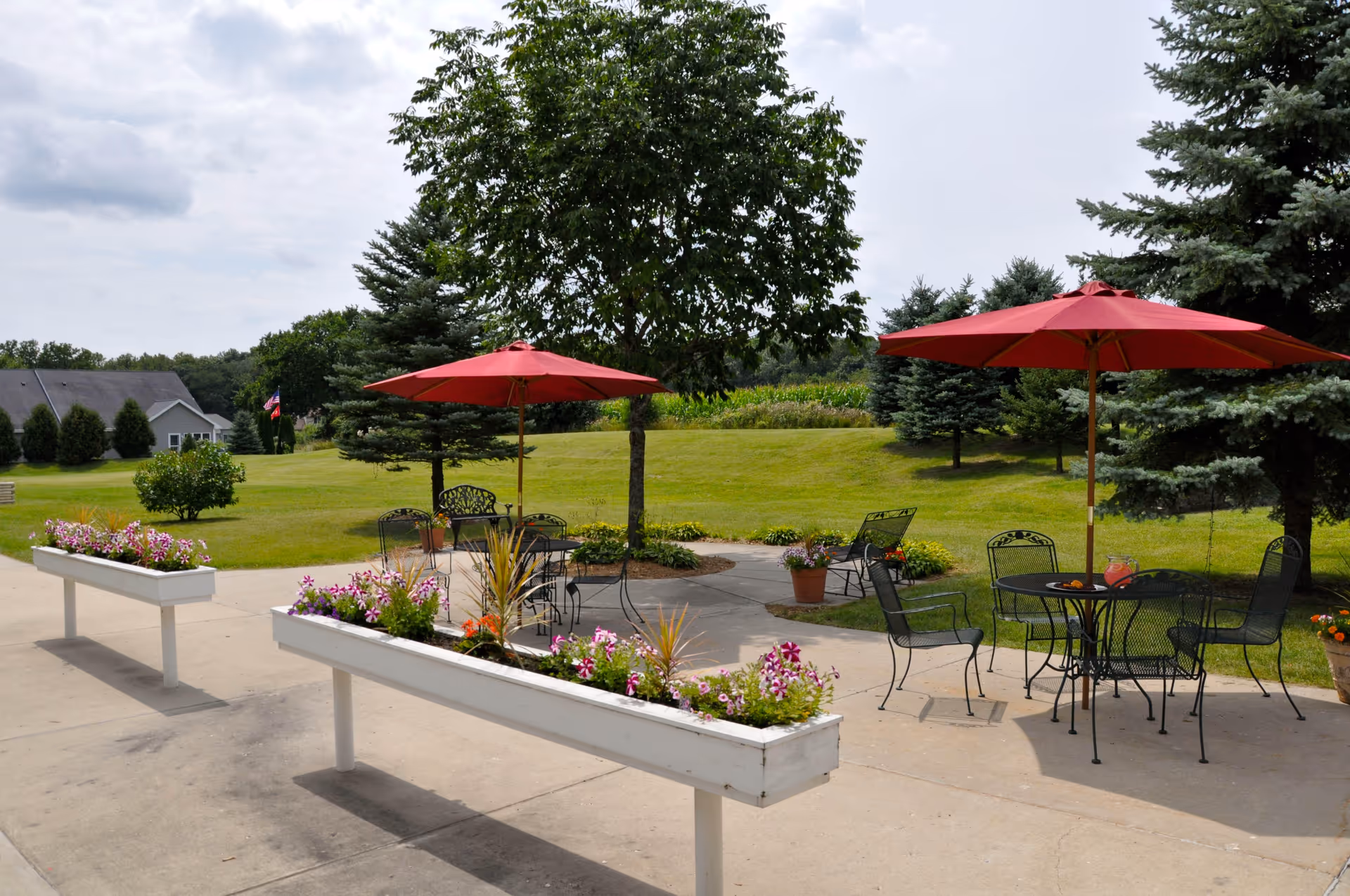 Outdoor patio area with metal tables and chairs under red umbrellas, surrounded by flower planters and green grass with trees in the background under a partly cloudy sky.
