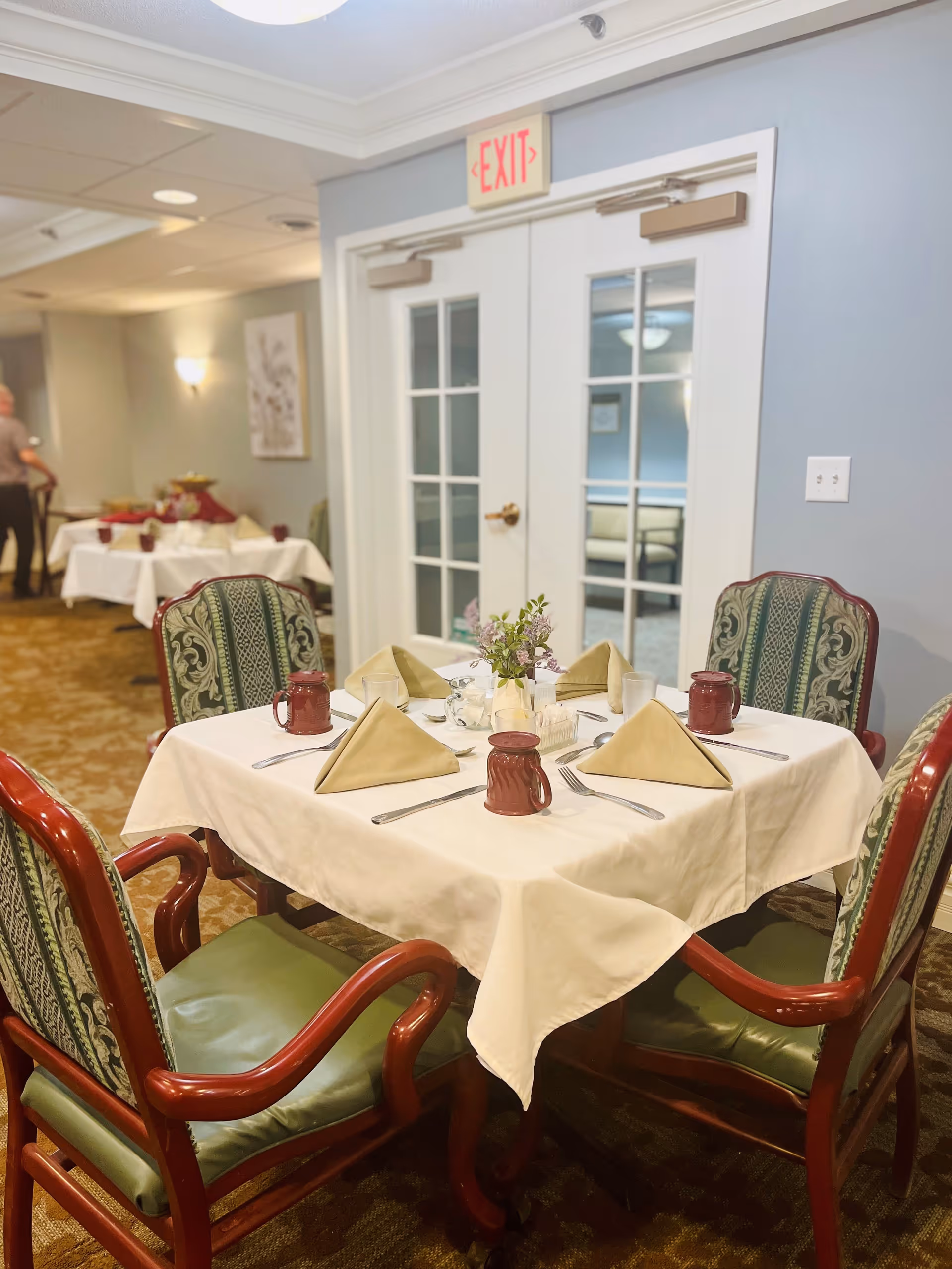 A dining table set for four with beige napkins folded in a triangle, red mugs, silverware, and a small flower arrangement in the center. The table is covered with a white tablecloth and surrounded by four wooden chairs with green cushioned seats and patterned backs. In the background, there is a buffet table with food and a person standing near it. Double glass doors with an exit sign above them are visible on the wall behind the table.
