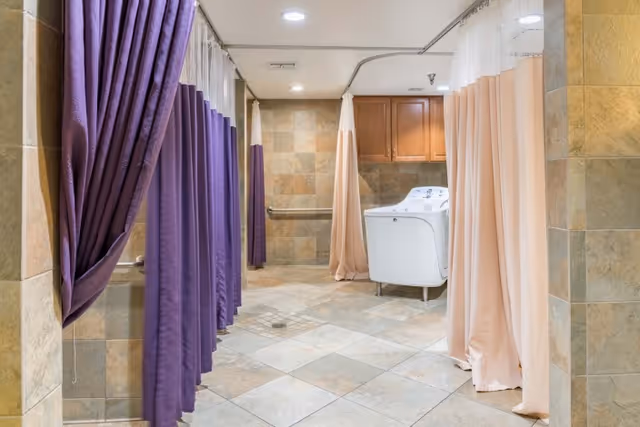 A tiled room with multiple shower stalls separated by purple and beige curtains, a white washing machine, and wooden cabinets mounted on the wall.
