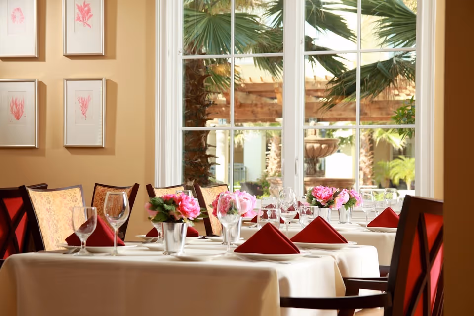 A dining room with tables set with white tablecloths, red folded napkins, wine glasses, and small flower arrangements in silver vases. The room has large windows showing palm trees and an outdoor fountain. The walls have framed botanical prints.