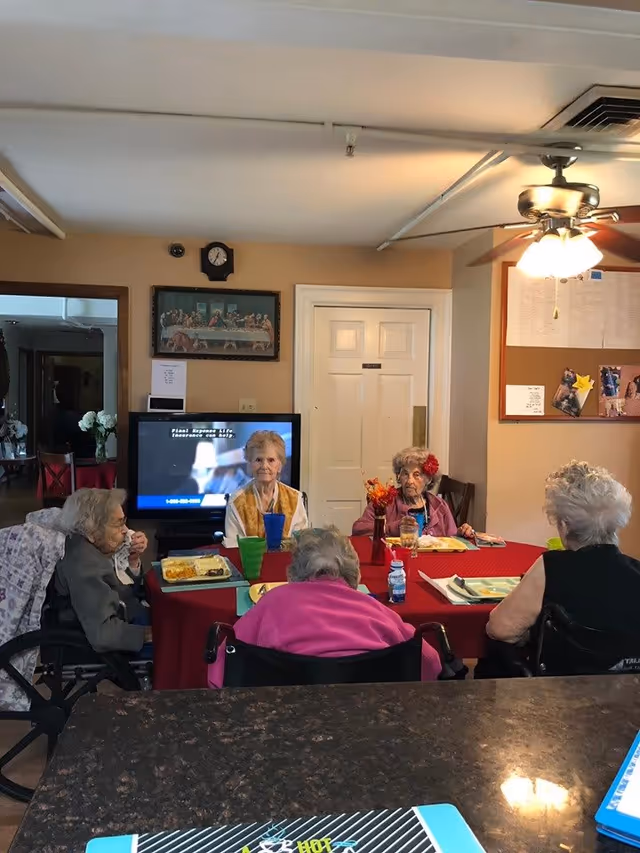 Five elderly women sitting around a rectangular table covered with a red tablecloth, having a meal together in a dining area. The room has beige walls, a ceiling fan with lights, a TV displaying a commercial, a clock, and a framed picture on the wall. Some of the women are in wheelchairs, and there are colorful cups and plates on the table.