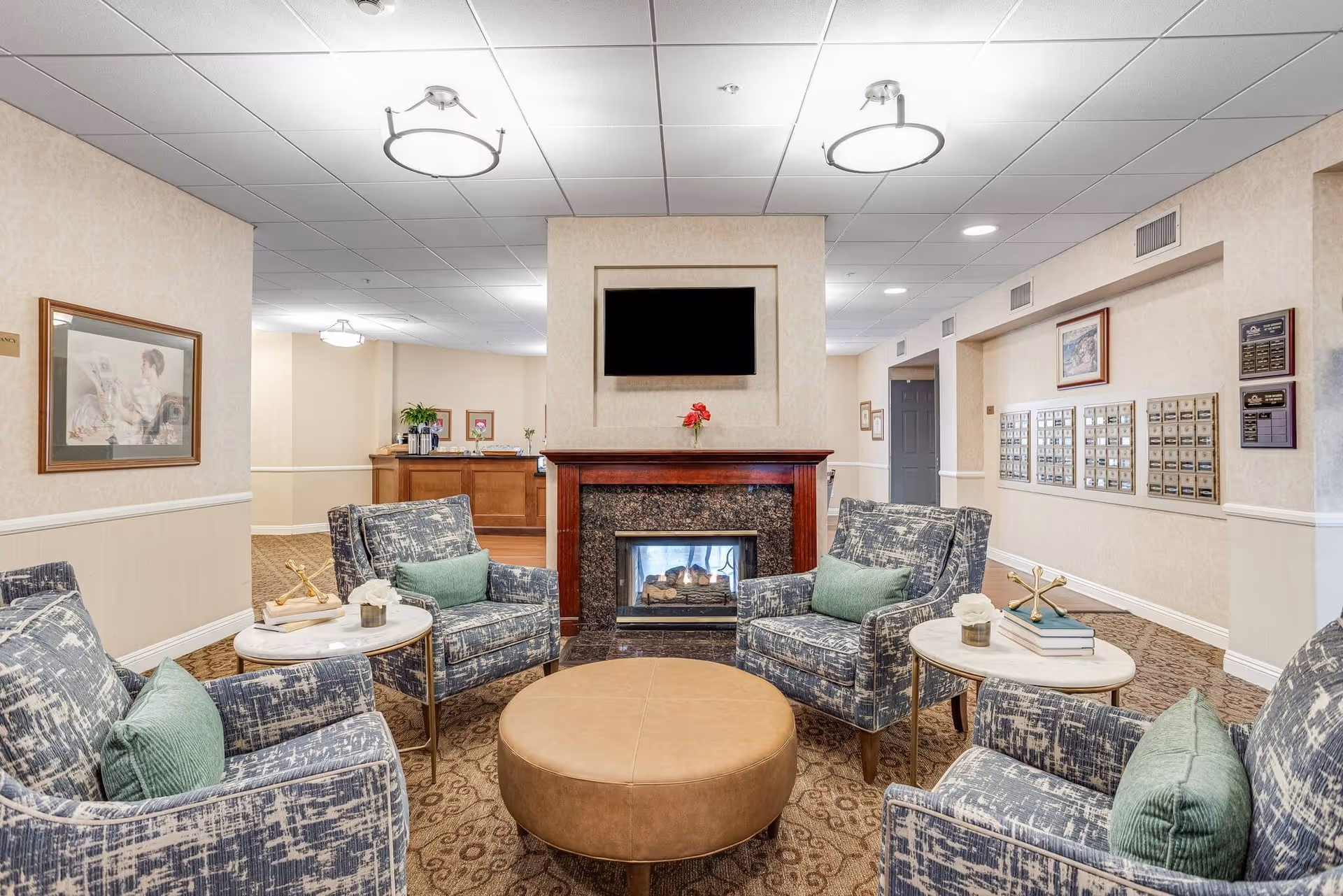 A cozy seating area in a senior living facility with four patterned armchairs arranged around a round leather ottoman. Each chair has a green cushion. There are two small round side tables with decorative items on them. In the background, there is a fireplace with a TV mounted above it and a reception desk further back. The walls are light-colored with framed artwork and a set of mailboxes on the right side.