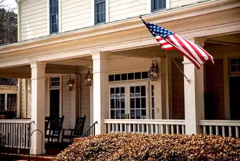 Front porch of a senior living facility with rocking chairs and an American flag.