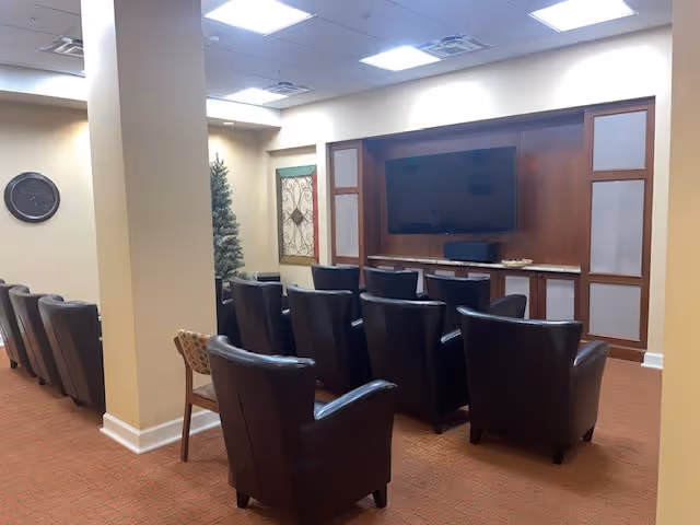 A small theater or media room with two rows of black leather chairs facing a large flat-screen TV mounted on a wooden wall unit. The room has beige walls, a carpeted floor, a clock on the wall, and a decorative framed piece next to a small artificial tree.
