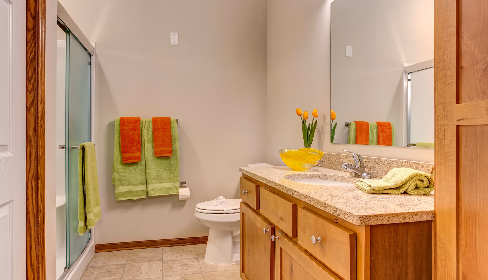 A clean and well-lit bathroom featuring a wooden vanity with a granite countertop, a sink with a faucet, a large mirror, and a vase with yellow tulips. There are green and orange towels hanging on the wall and a toilet next to the vanity. A glass shower door is visible on the left side.