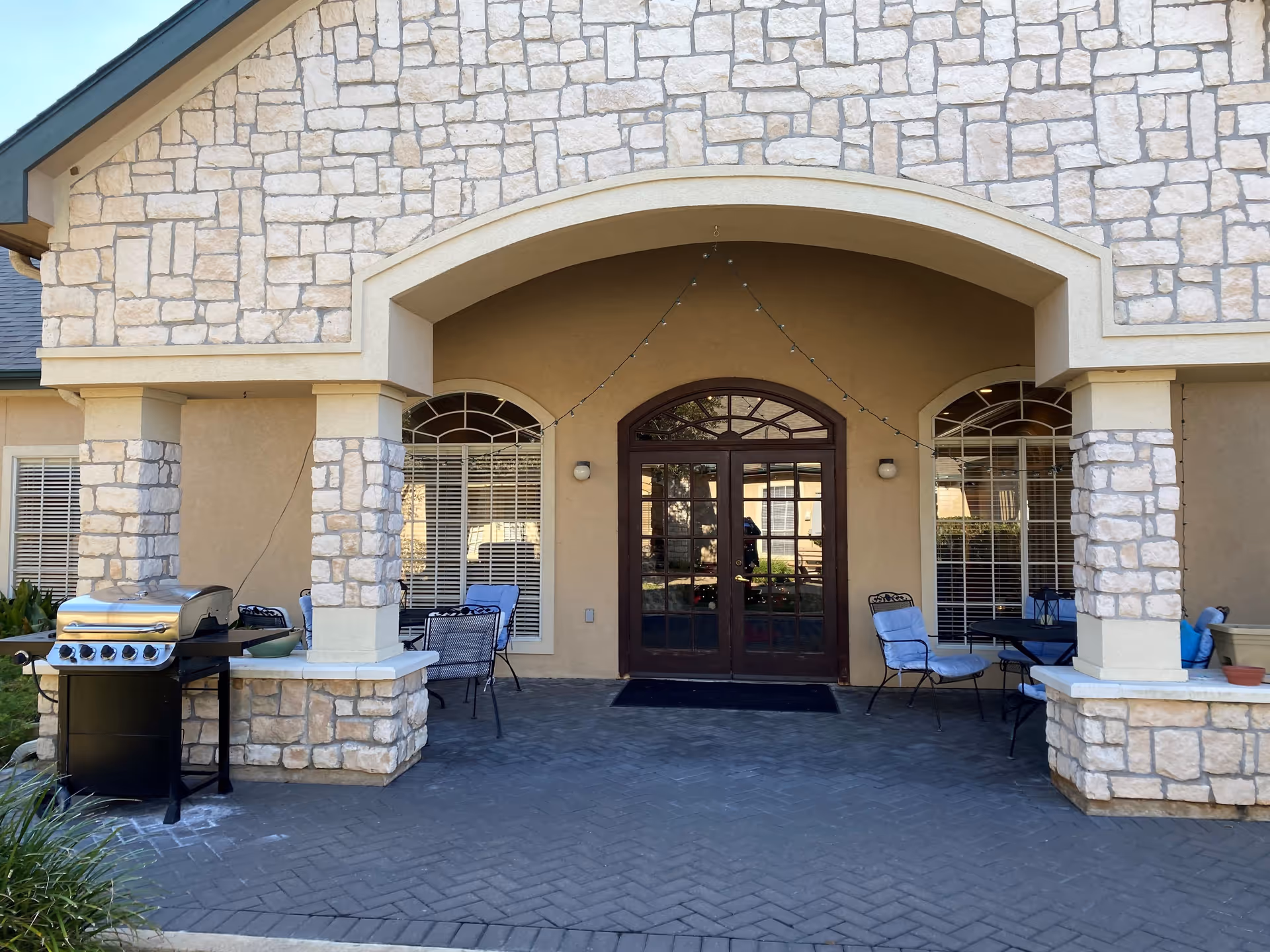 Outdoor covered patio area with stone pillars and beige walls. There are two sets of windows with white blinds on either side of a double glass door entrance. Several cushioned chairs and tables are arranged under the patio. A stainless steel grill is positioned on the left side near one of the stone pillars.