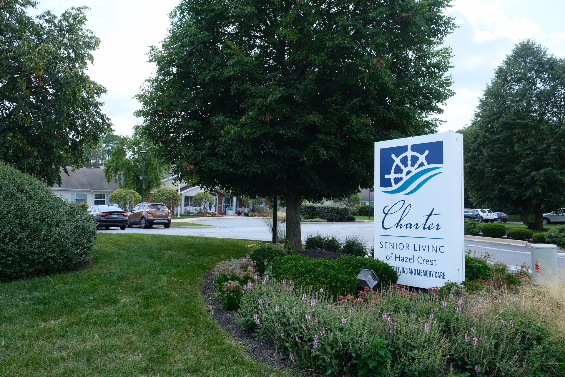 Outdoor view of Charter Senior Living of Hazel Crest facility sign surrounded by green bushes, flowers, and trees with parked cars and residential-style buildings in the background under a cloudy sky.