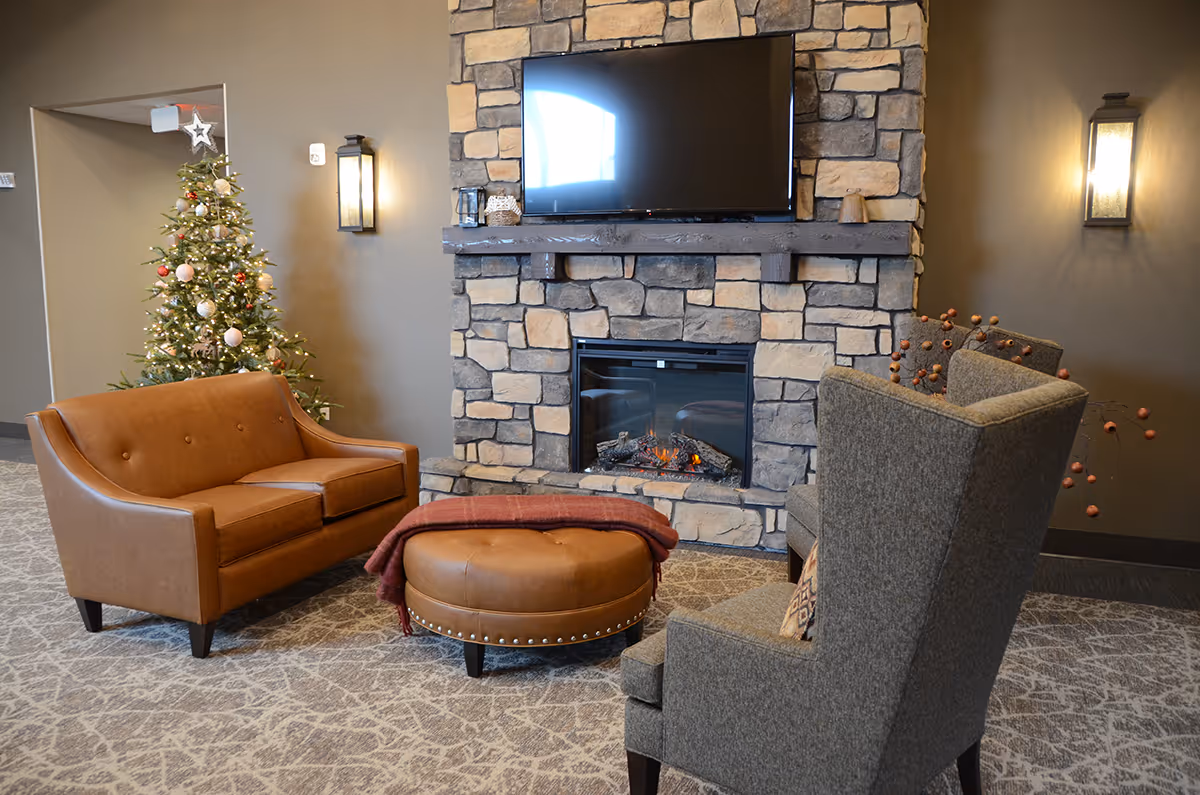 Cozy living room area with a stone fireplace, a mounted flat-screen TV above it, a brown leather loveseat, a round leather ottoman with a red blanket, a gray upholstered armchair, and a decorated Christmas tree in the corner.