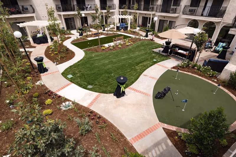 A landscaped outdoor courtyard area in a senior living facility featuring green lawns, a putting green with golf clubs and balls, paved walkways with red brick accents, benches, small trees and plants, and a shaded seating area with an umbrella. The courtyard is surrounded by a multi-story building with balconies overlooking the space.
