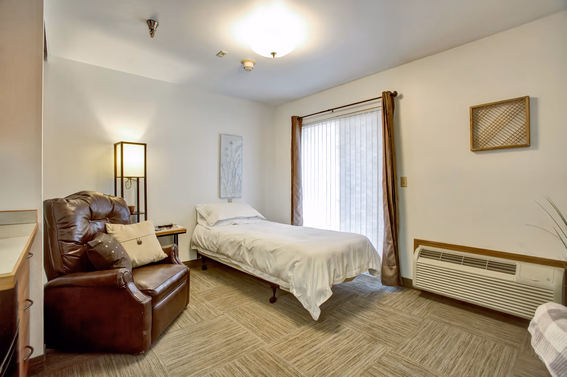 A senior living facility bedroom with a single bed covered in white bedding, a brown leather armchair with pillows, a floor lamp, a small side table, a large window with vertical blinds and brown curtains, a wall-mounted heater, and simple wall decorations.