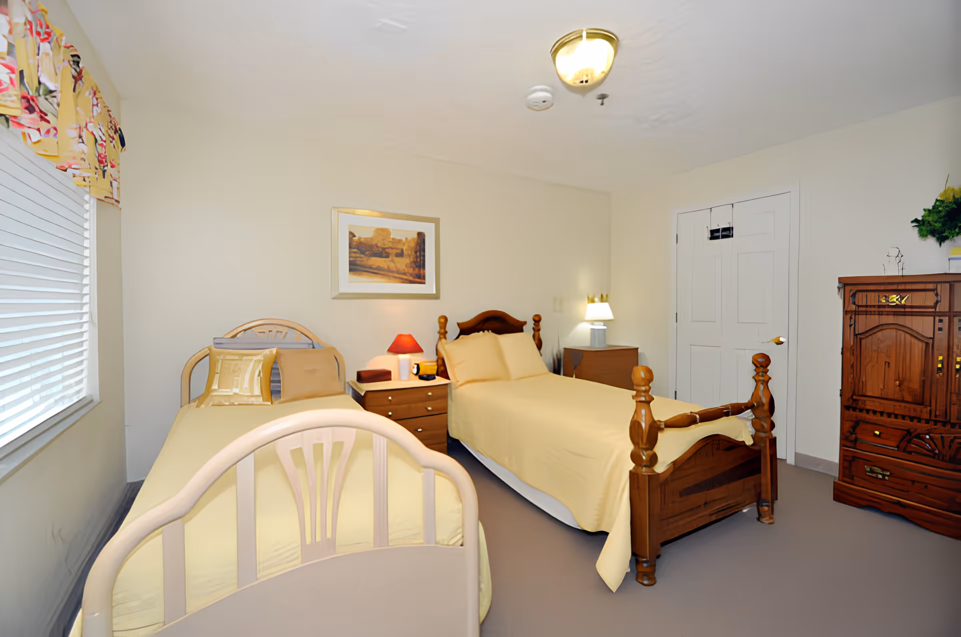 A bedroom with two single beds, each with yellow bedding. Between the beds is a wooden nightstand with a red lamp and other small items. There is a framed picture on the wall above the nightstand. The room has a window with blinds and a colorful valance on the left side. On the right side, there is a wooden dresser with a plant on top and a closed white door. The walls are light-colored and the floor is carpeted.