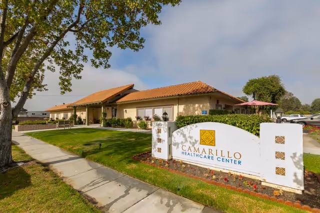 Exterior view of Camarillo Healthcare Center building with a tiled roof, surrounded by green grass, trees, and a sidewalk. A white sign with the facility's name is prominently displayed in the foreground.
