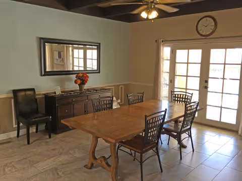 A dining room with a wooden table and six chairs, a sideboard with a vase of flowers, a large mirror on the wall, a ceiling fan with lights, and French doors with curtains leading outside.