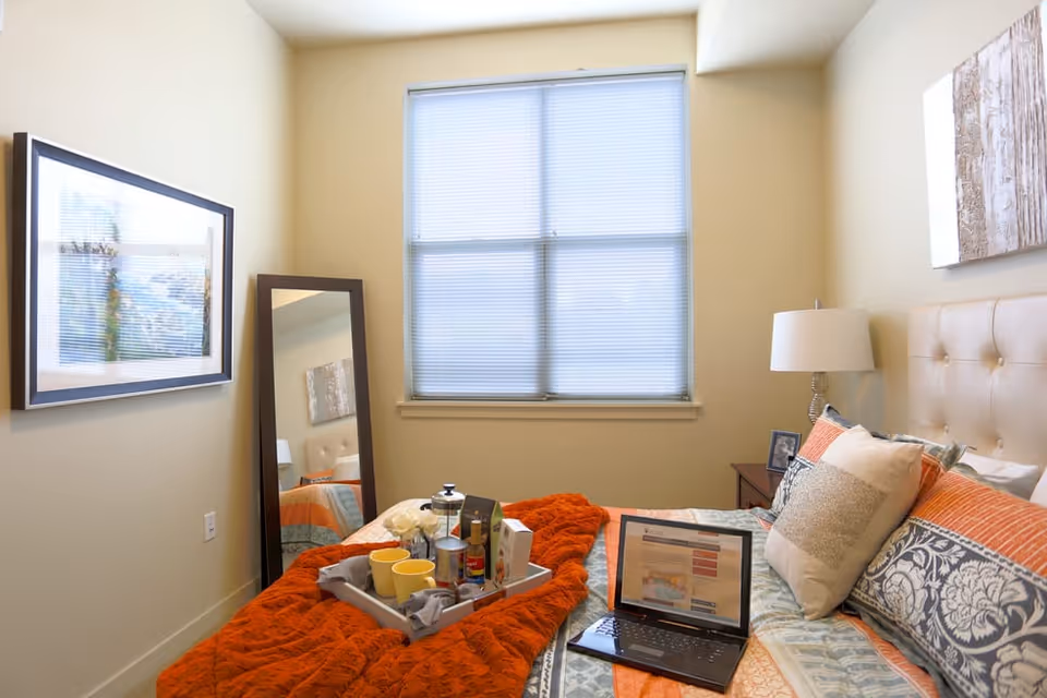 A cozy bedroom with a bed covered in a patterned quilt and an orange throw blanket. On the bed is a tray with cups, a teapot, and other items, along with an open laptop. The room has a large window with blinds, a tall mirror leaning against the wall, a framed picture on the wall, and a bedside table with a lamp and a photo frame.